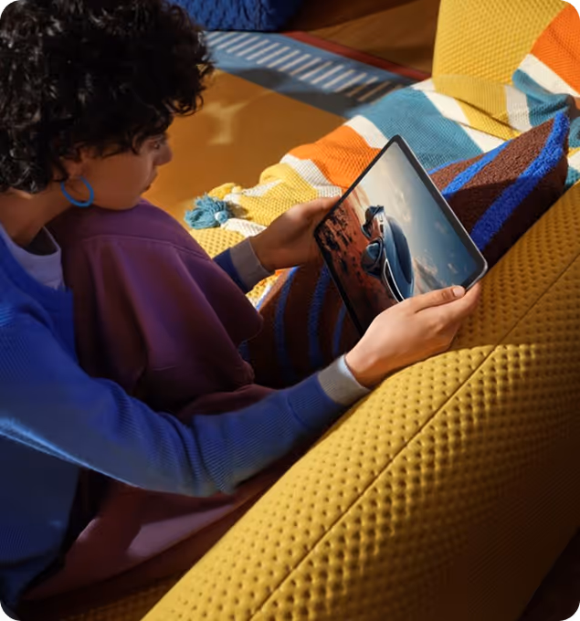 Person sitting on a yellow couch holding a tablet displaying an image of a car in a desert landscape.